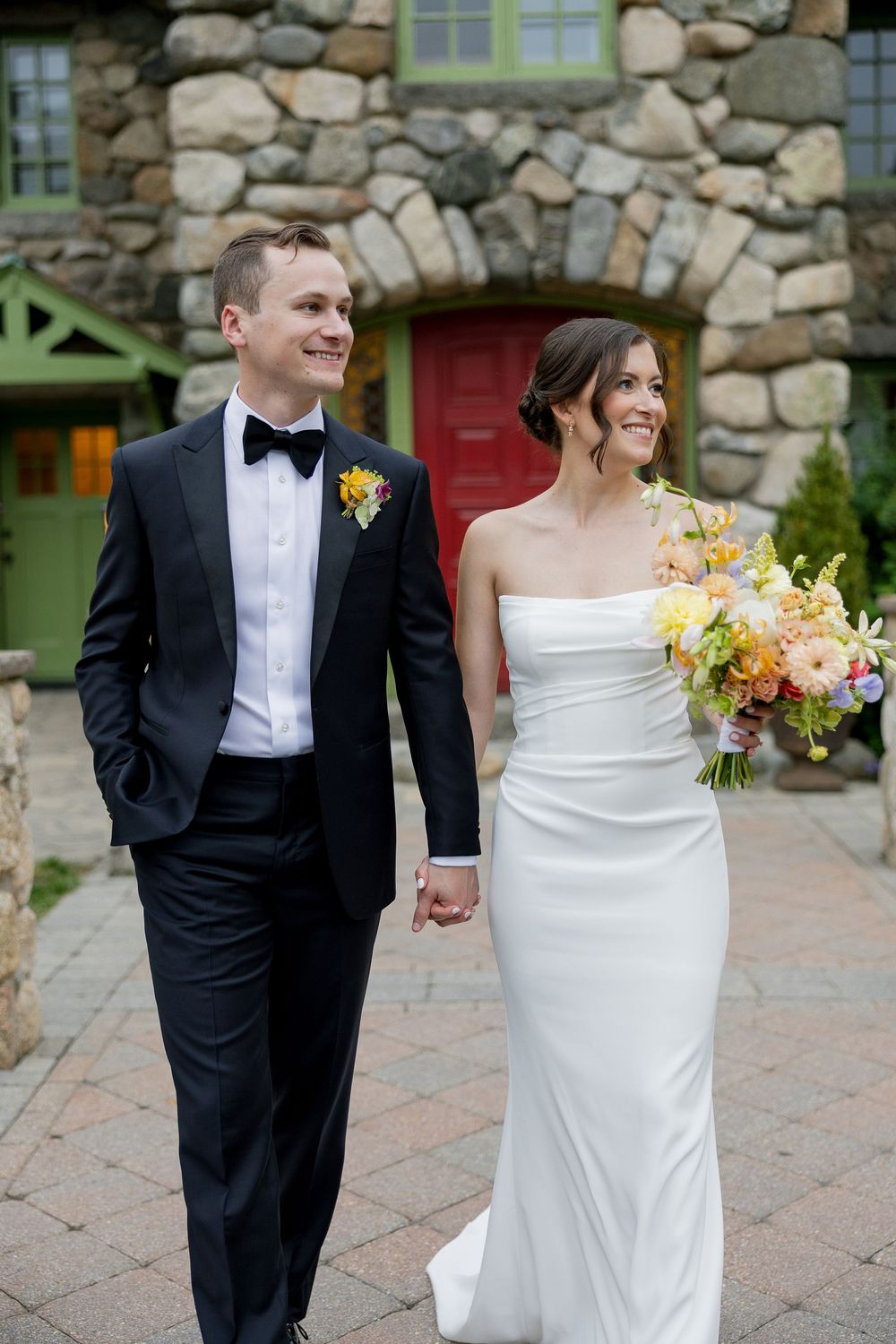 Smiling bride and groom in black tuxedo walking hand in hand outside rustic stone wedding venue.