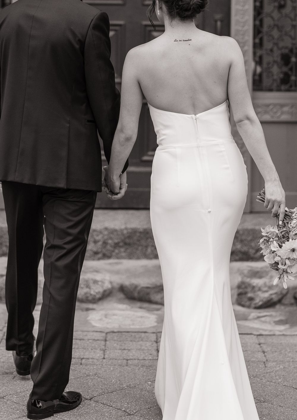 Black and white photo of bride in strapless fitted gown holding bouquet, walking hand in hand with groom.