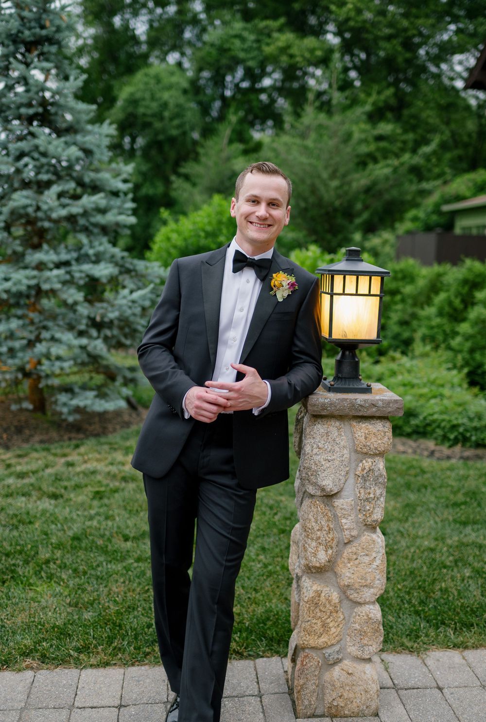 Smiling groom in black tuxedo leans against stone pillar lamp post surrounded by lush green garden.