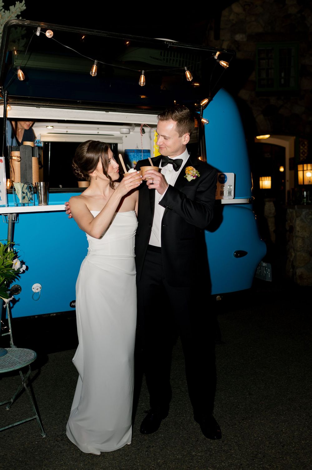 Bride in white gown and groom in black tuxedo share a toast in front of a vintage blue food truck at night.