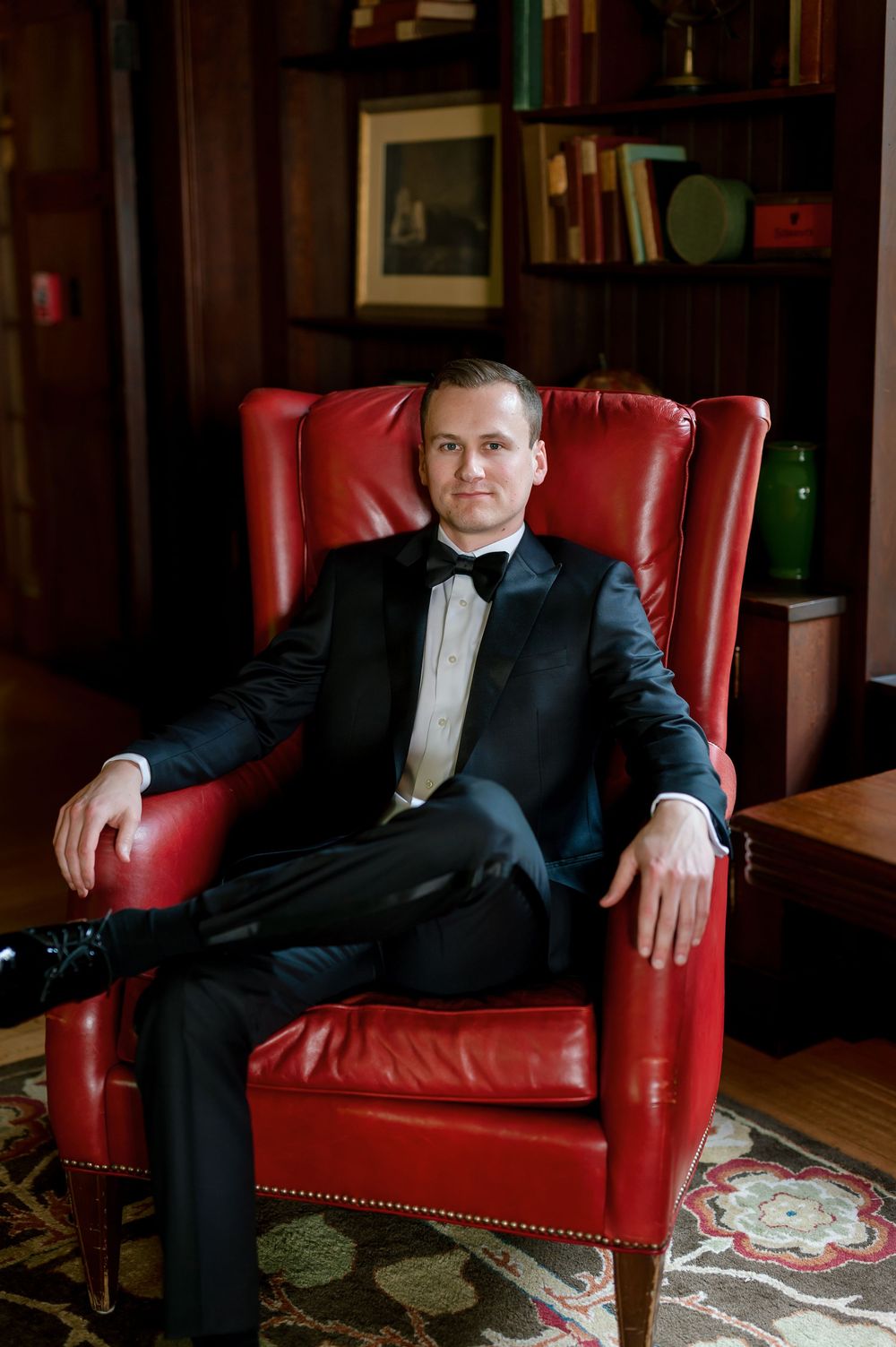 A man in a tuxedo sits confidently in a red leather wingback chair in an elegant wood-paneled library.