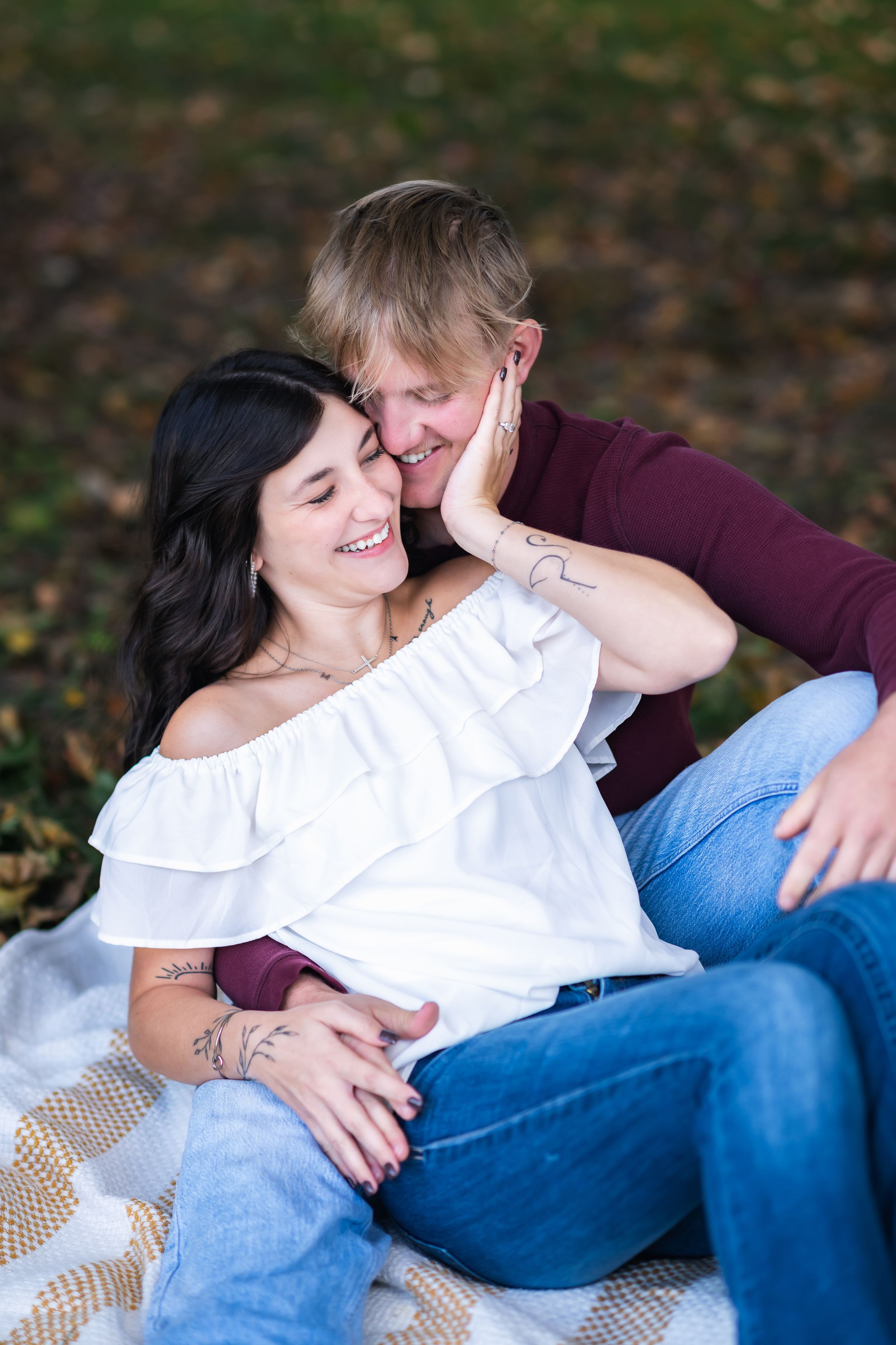 Couple sitting on blanket outdoors, woman in white off-shoulder top smiling as man in burgundy shirt kisses her cheek.