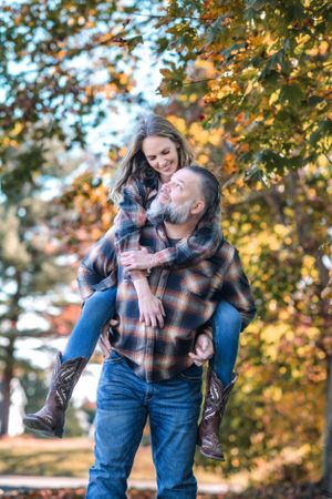 Playful piggyback ride moment between couple wearing matching flannel shirts in fall foliage.