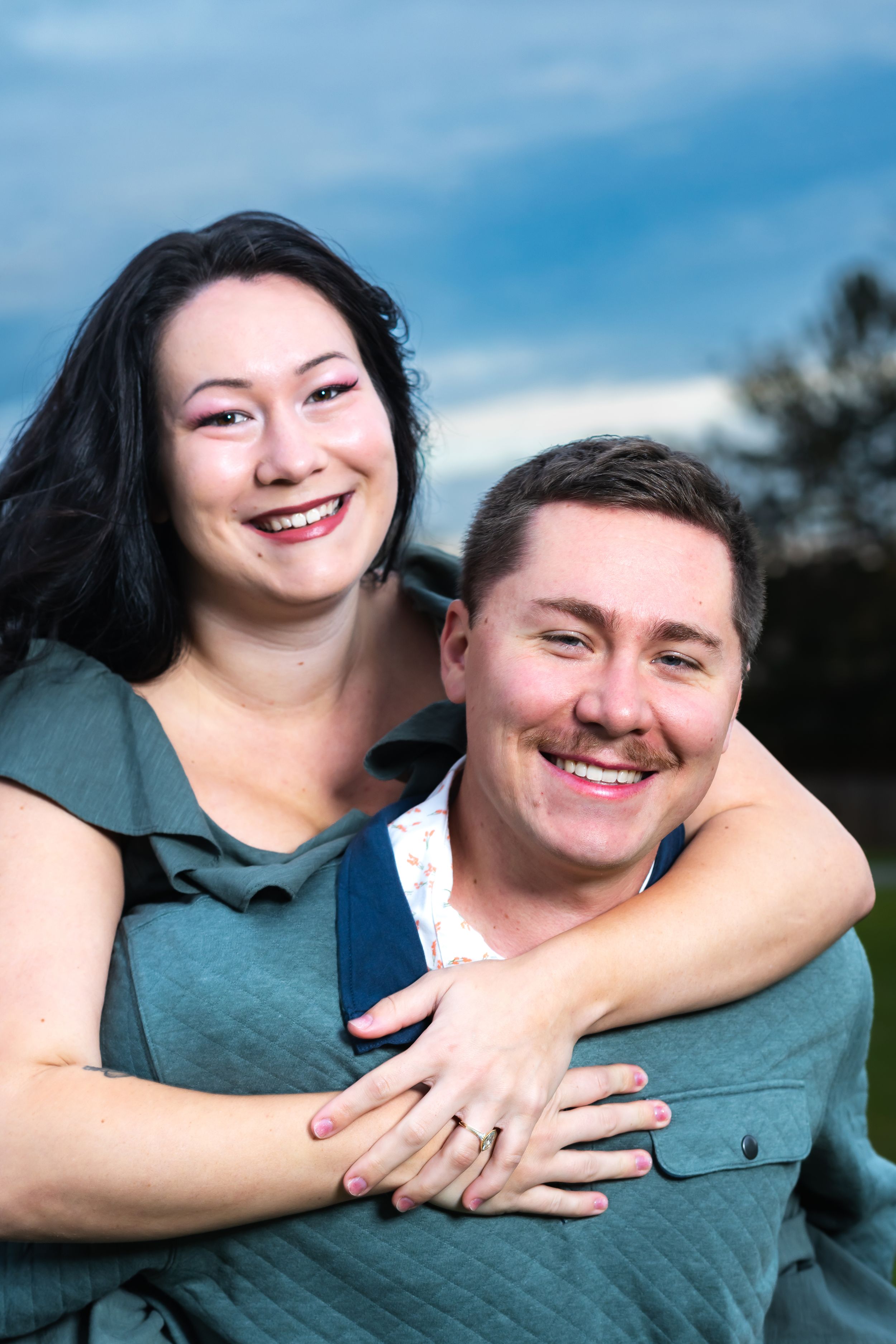 Happy couple smiling outdoors, woman hugging man from behind, wearing teal outfits against a blue sky backdrop.