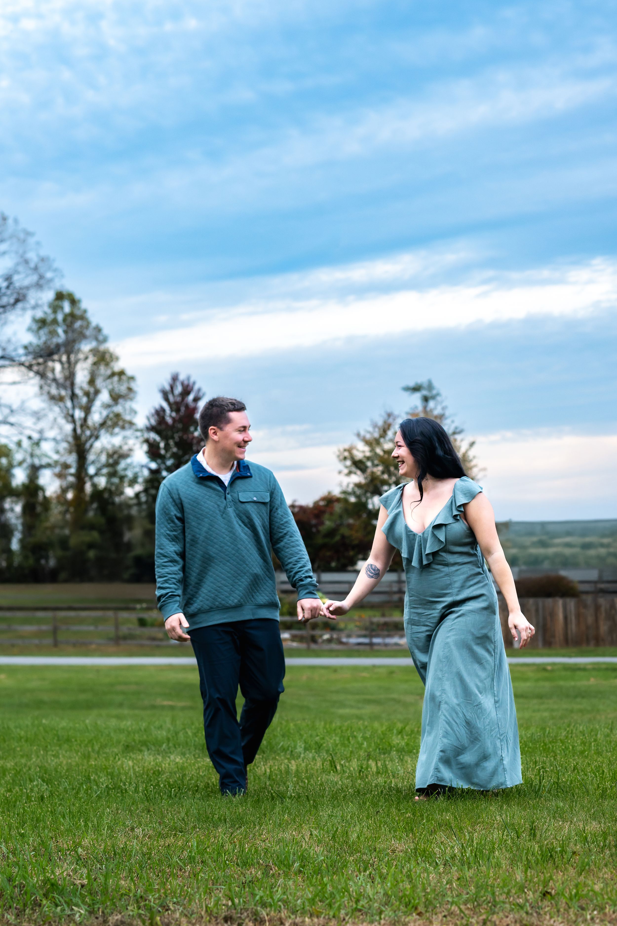A couple holding hands and walking on green grass, smiling at each other, dressed in matching teal outfits outdoors.