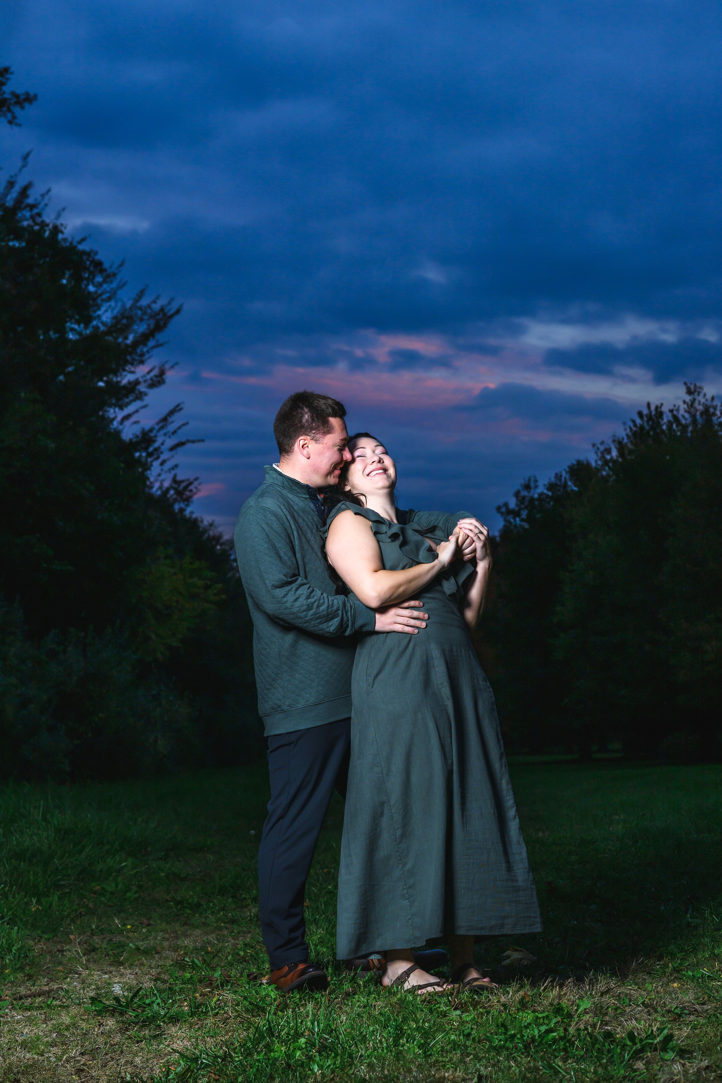 Couple embracing at dusk in a lush green field, wearing matching olive outfits under a dramatic purple and blue sky.