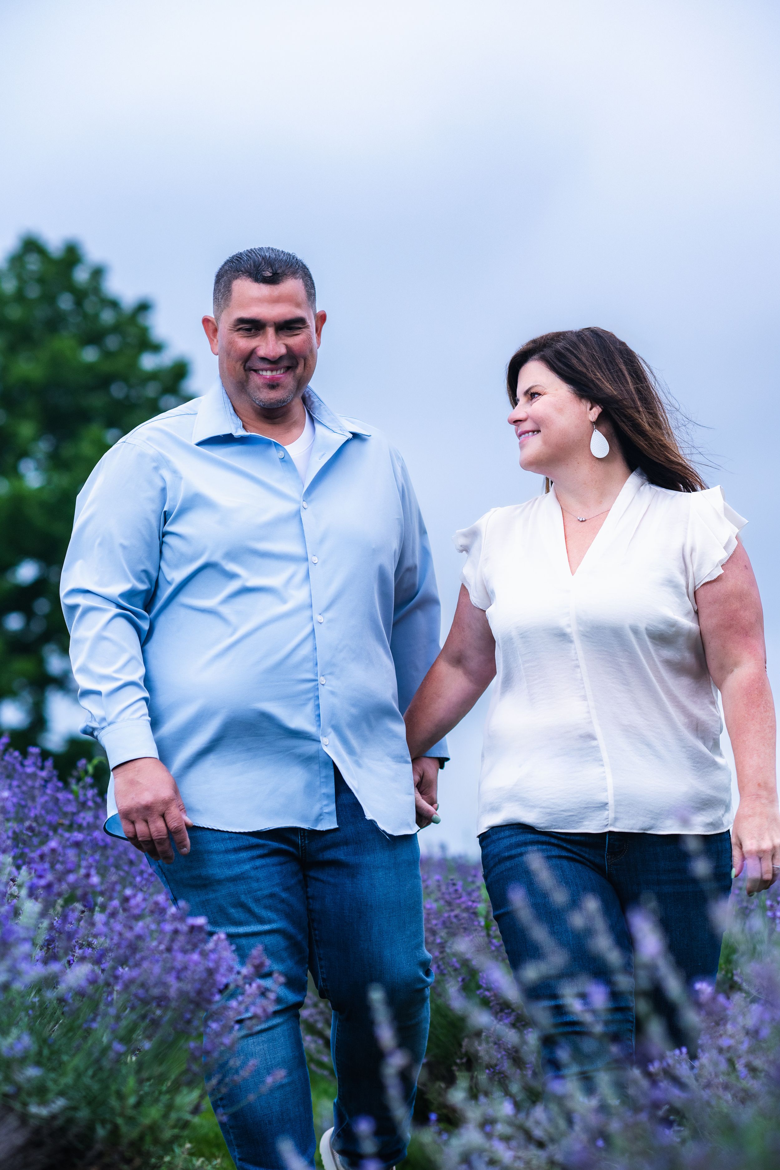 Couple holding hands and walking through a blooming lavender field, smiling at each other on a cloudy day.