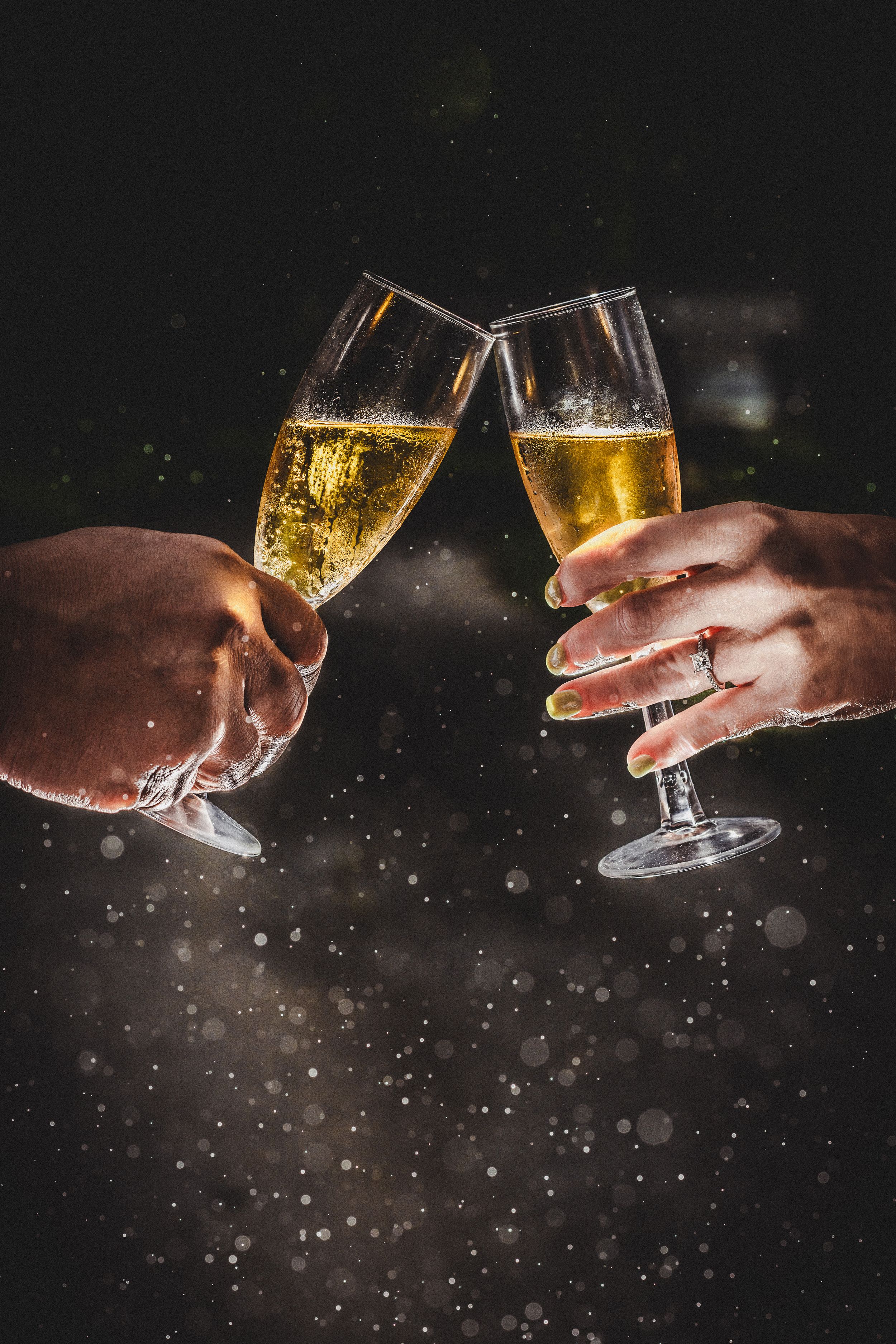 Two hands clinking champagne flutes in a celebratory toast against a dark bokeh background.