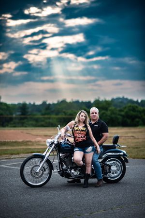 Couple poses on motorcycle against dramatic cloudy sky in outdoor parking lot.