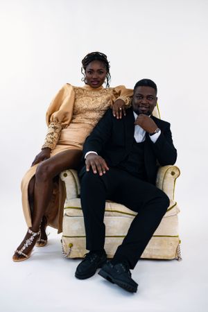 Elegant couple poses on cream vintage chair wearing formal gold dress and black suit.