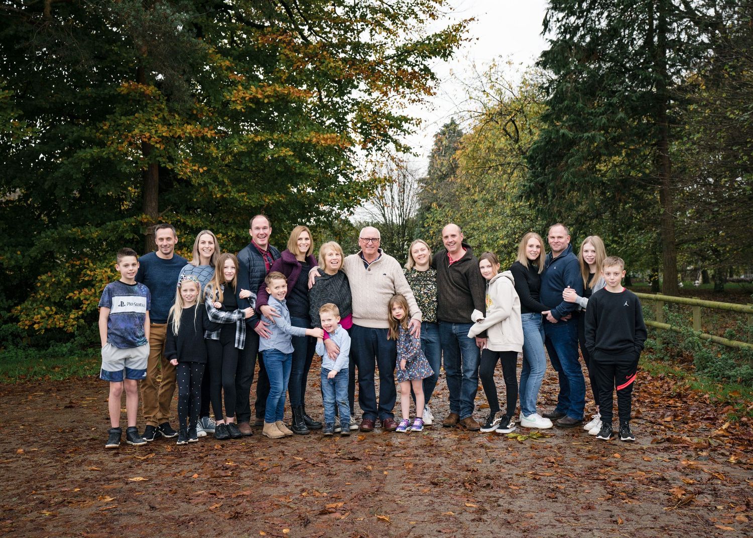Large multigenerational family group portrait outdoors in autumn park with fallen leaves and evergreen trees.