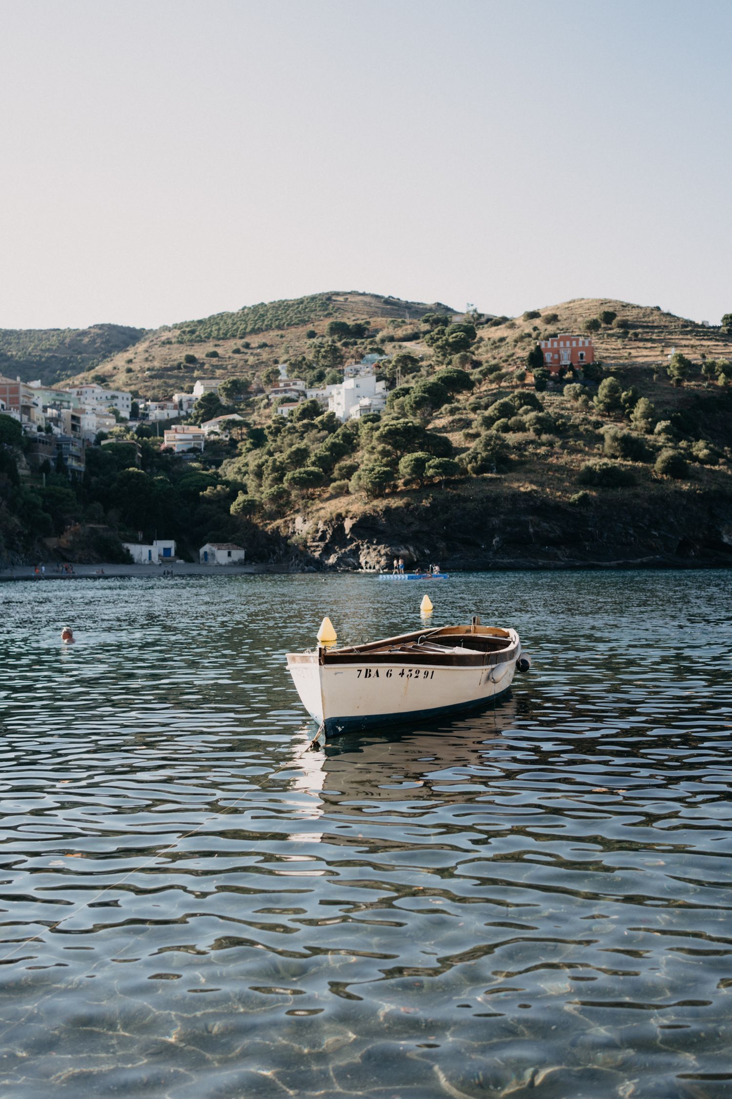 Sunbathers on a rocky Mediterranean beach with lush green trees and elegant villas on a cliff above the shoreline.
