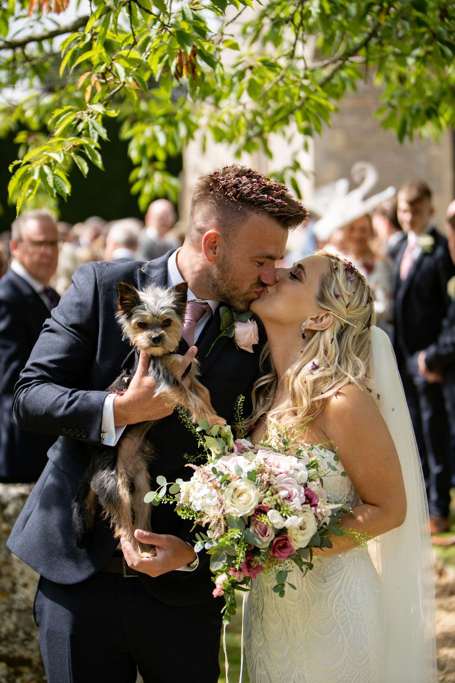 Bride and groom share a kiss holding a Yorkshire Terrier puppy, bride holds a pink floral bouquet at outdoor wedding.