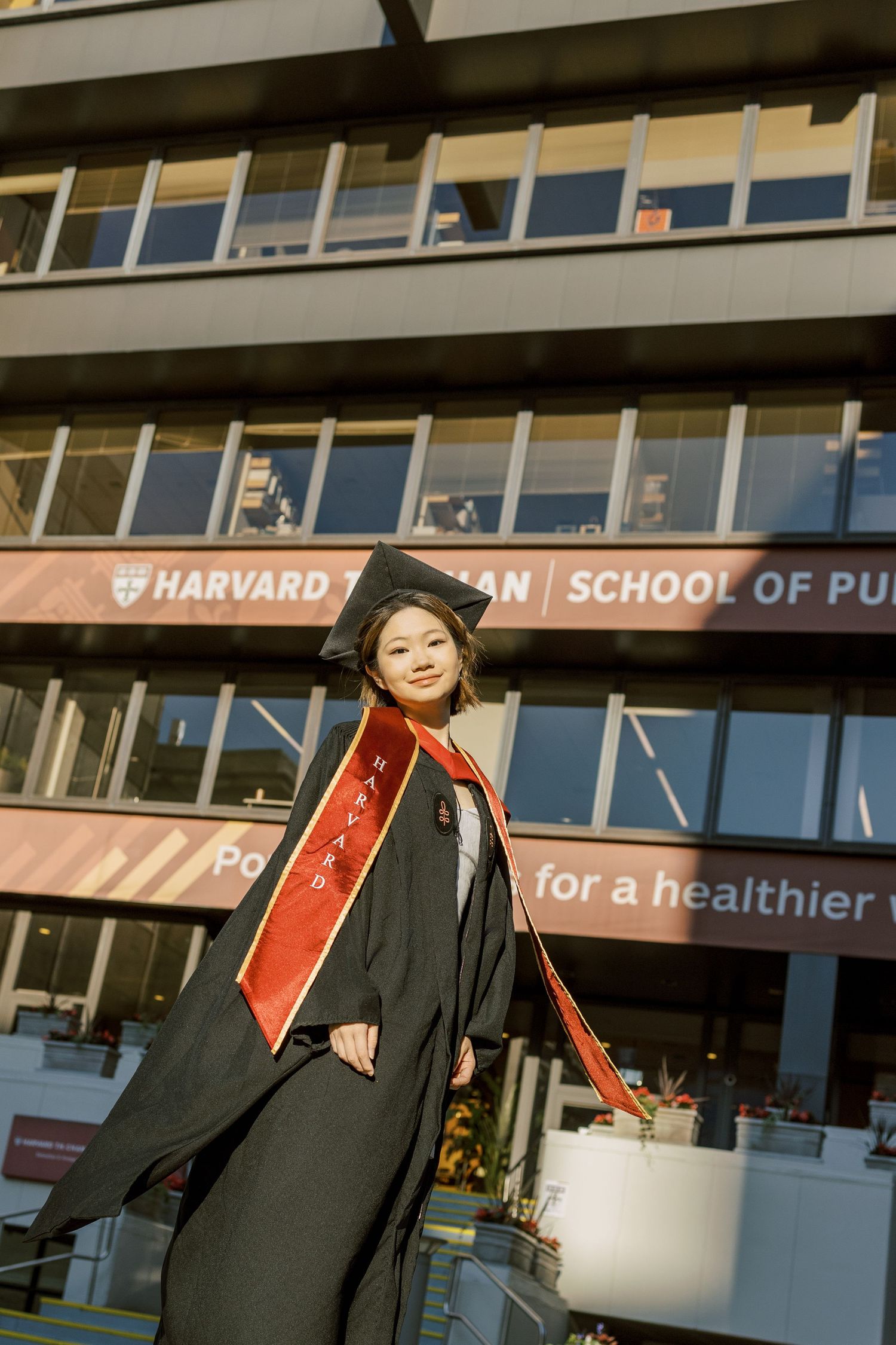 Harvard Chan School of Public Health graduate poses proudly in black cap and gown with red sash outside campus building.