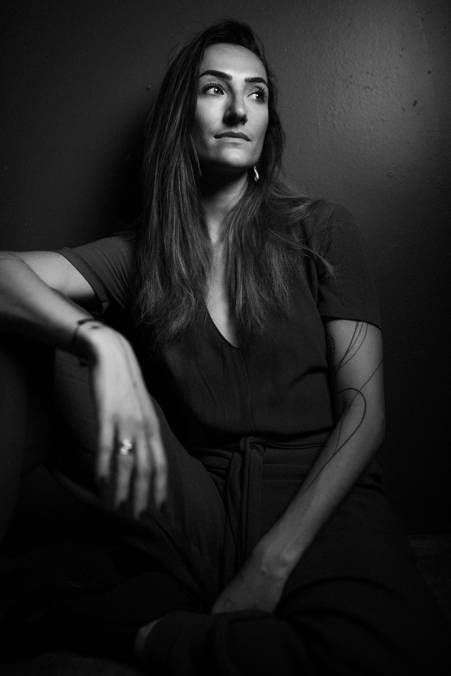 Black and white portrait of a woman in dark outfit sitting against a wall, gazing upward thoughtfully.