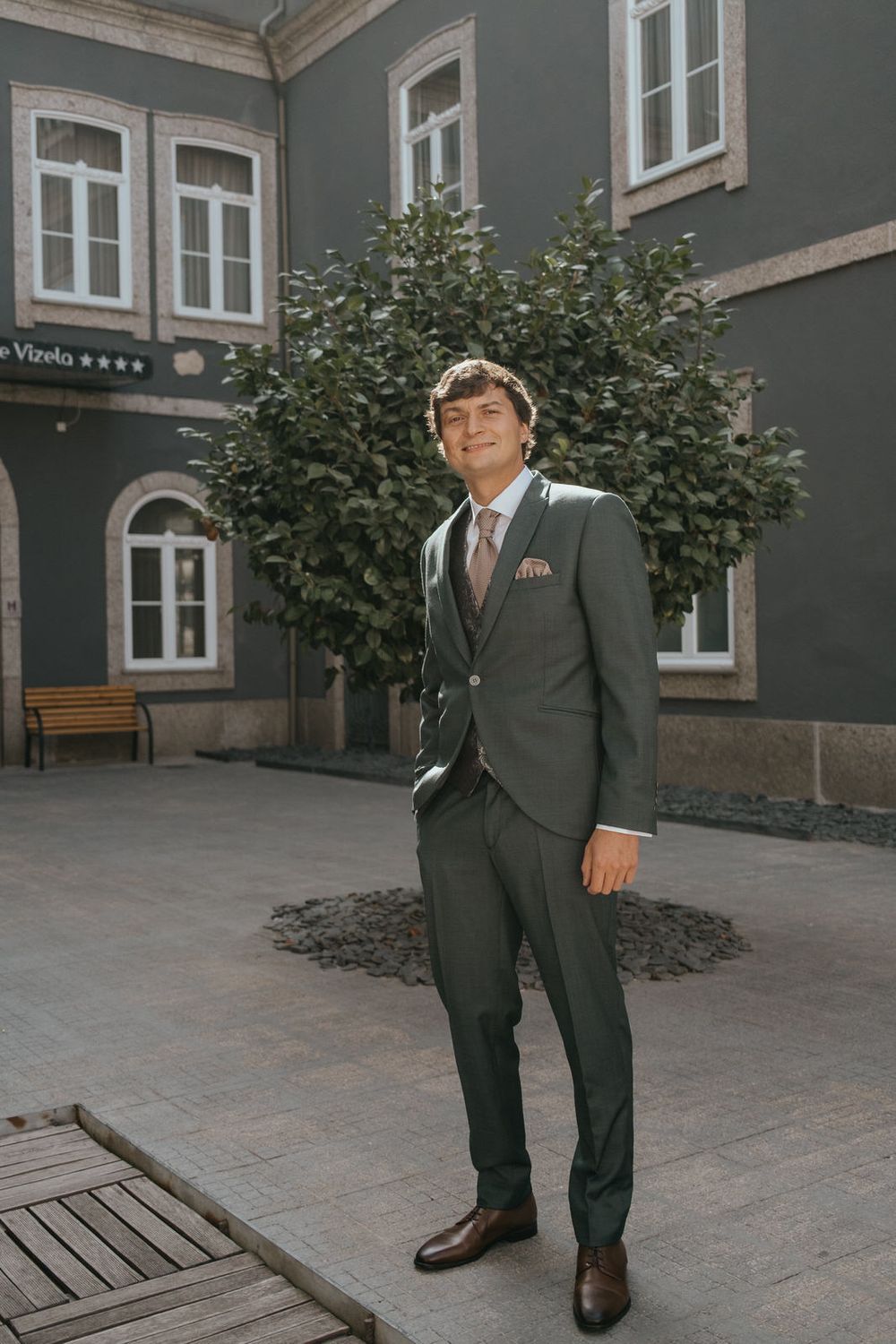 Young man in a tailored gray suit with tie stands confidently in a European courtyard with lush trees and elegant building.