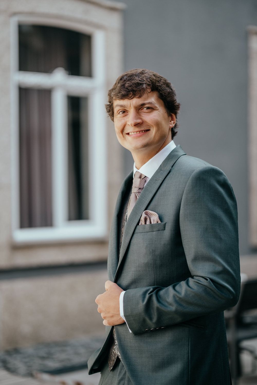 Smiling young man in a gray suit with pink tie and pocket square posing outdoors near a building.