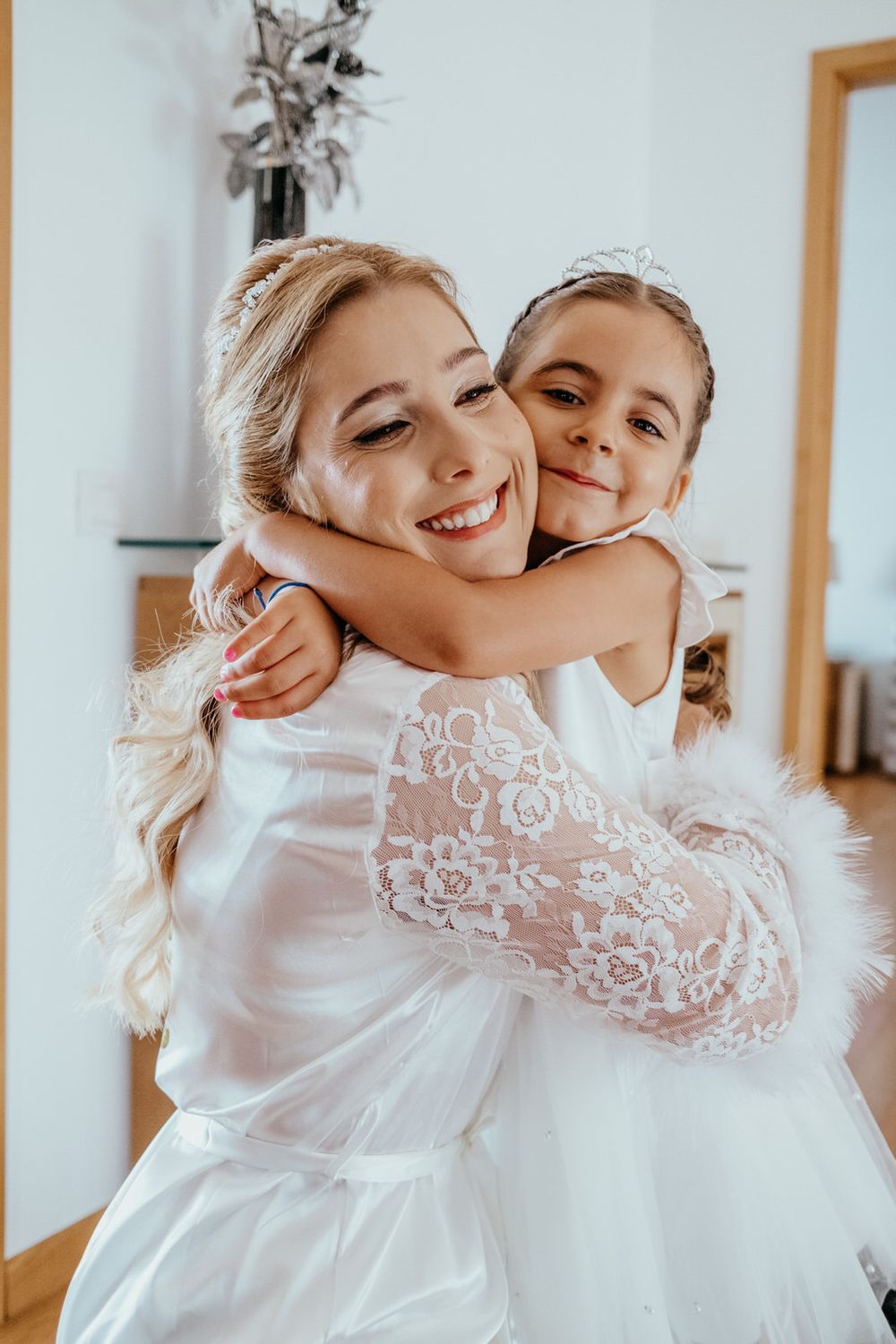 Bride in white lace dress hugging a young flower girl, both smiling warmly in a bright indoor setting.