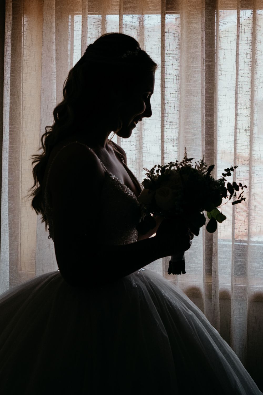 Silhouette of a bride in a ball gown holding a floral bouquet, backlit by sheer curtains.