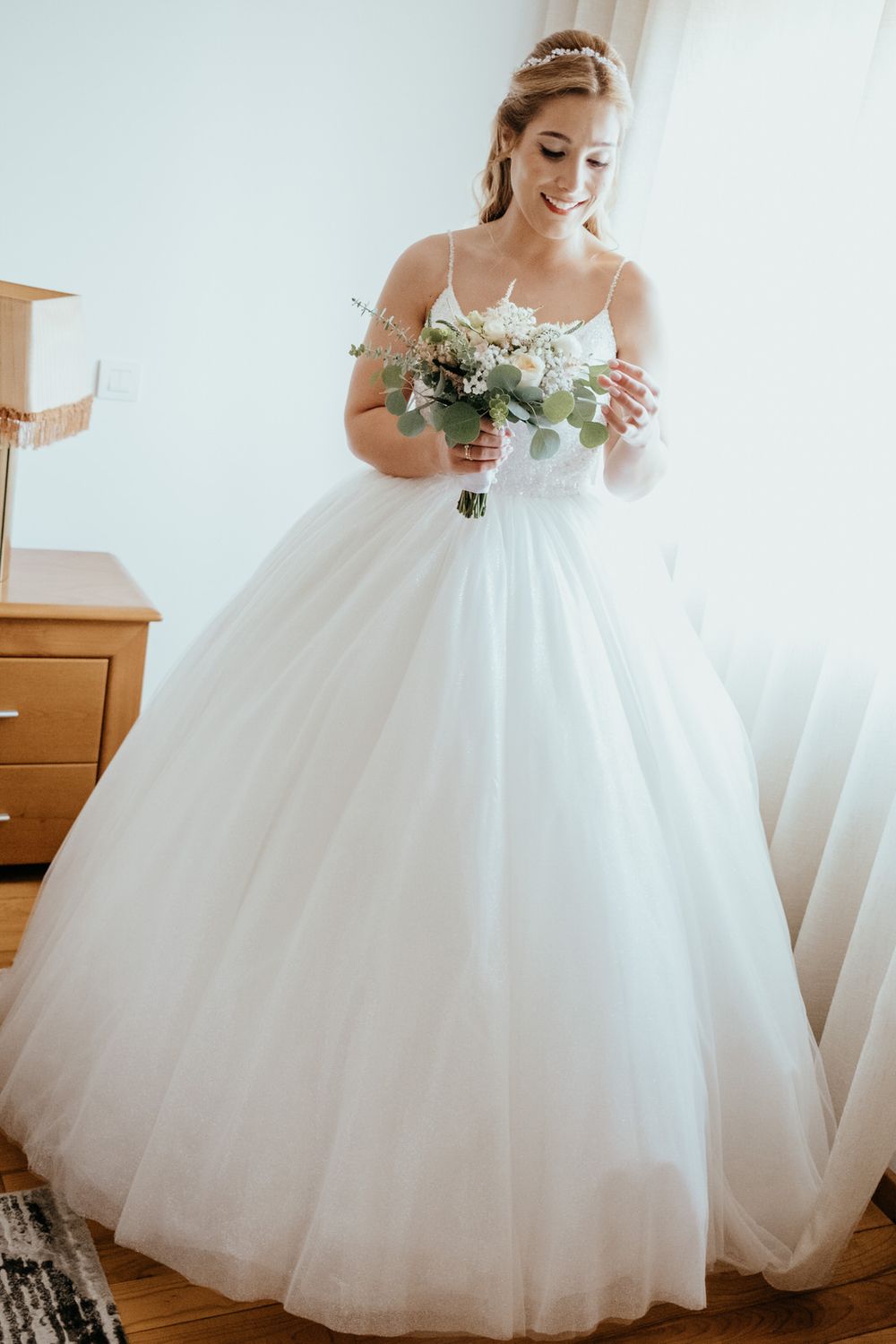 Smiling bride in a white ball gown holding a bouquet of white flowers and greenery, standing by a window.