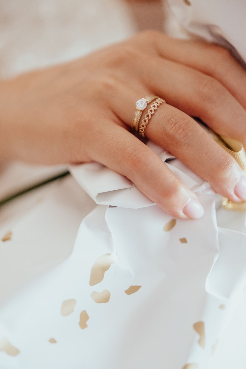 Close-up of a woman's hand wearing a diamond solitaire engagement ring and gold band on white fabric.