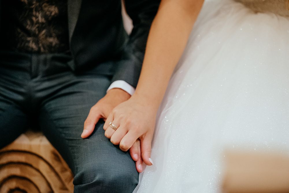 Bride and groom holding hands showing wedding ring, wearing white dress and grey suit at wedding ceremony.