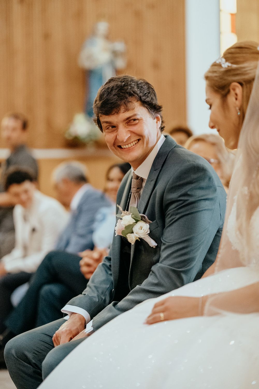 Smiling groom in gray suit with boutonniere sits beside bride in white dress during church wedding ceremony.