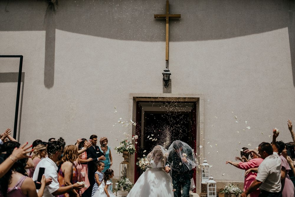Bride and groom exit church as guests toss rice, with a gold cross above the doorway.