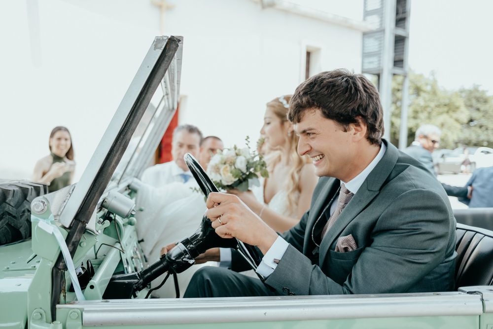 Smiling groom in gray suit driving vintage convertible car with bride holding bouquet at outdoor wedding.