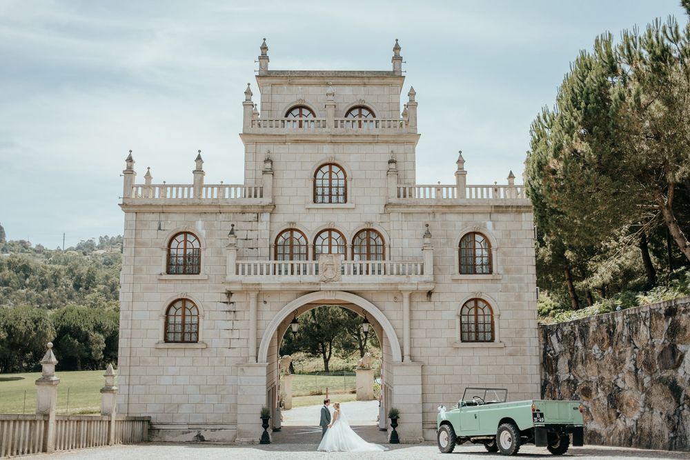 Bride and groom stand beneath ornate stone arch gateway of elegant palace venue with vintage green Land Rover.