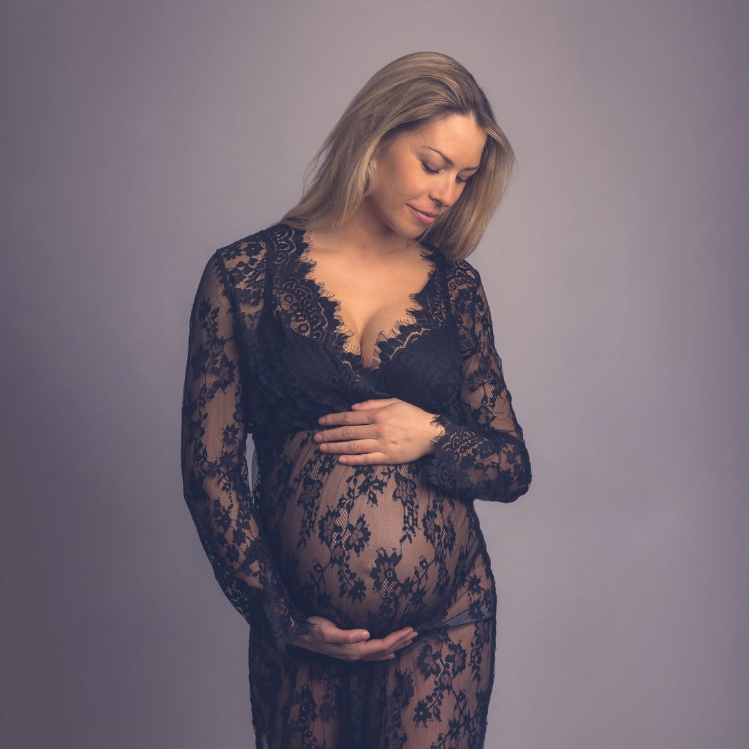Pregnant woman in black lace maternity gown cradling her baby bump during an elegant studio maternity photoshoot.