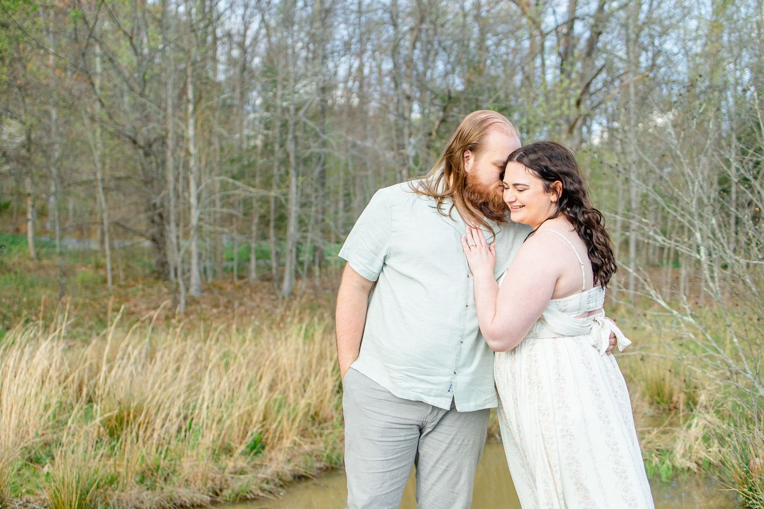 Couple sharing a tender moment outdoors, woman in white dress leaning on bearded man in mint shirt, surrounded by bare trees.