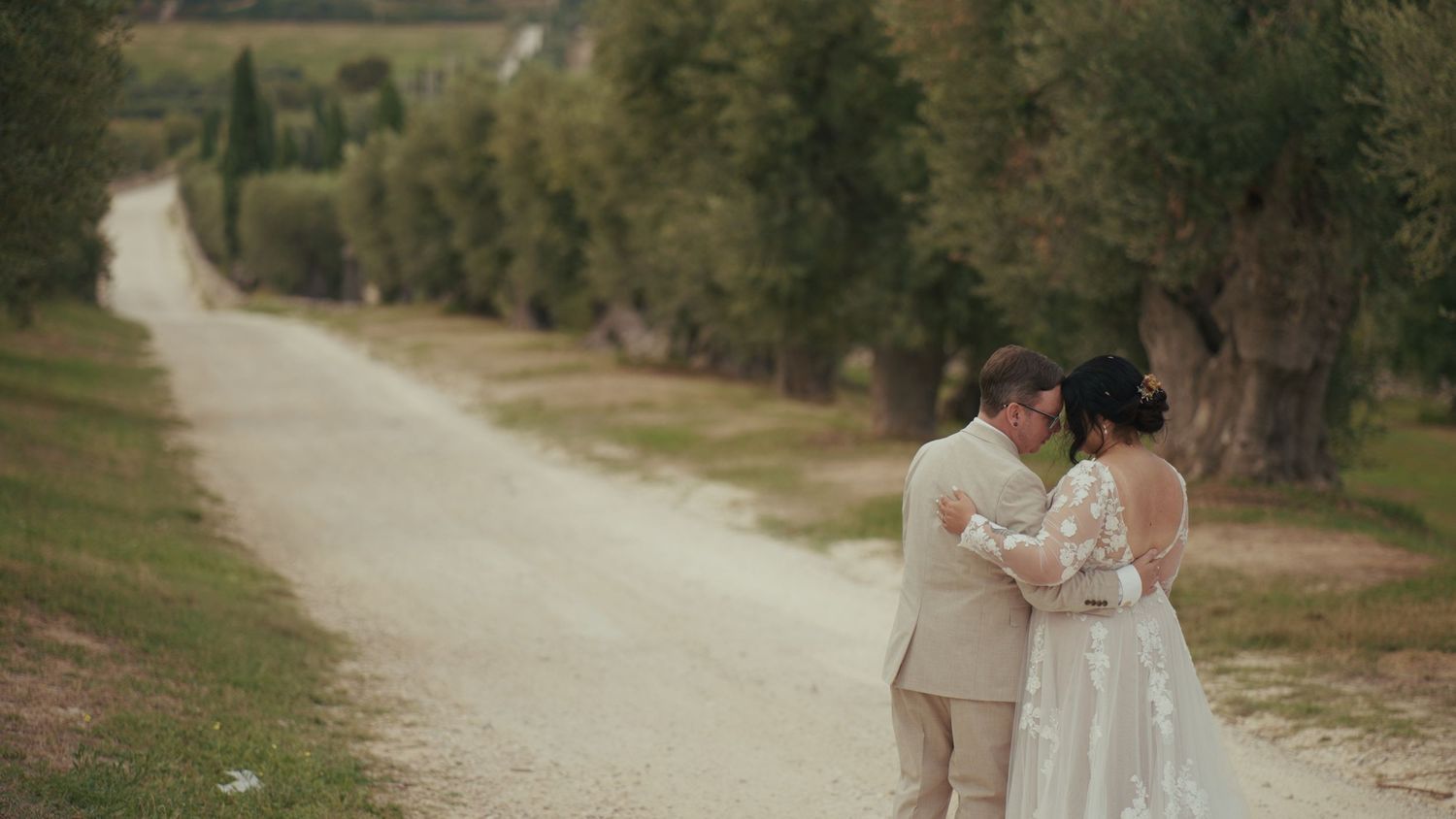 A bride and groom embrace on a scenic rural dirt road lined with trees on their wedding day.