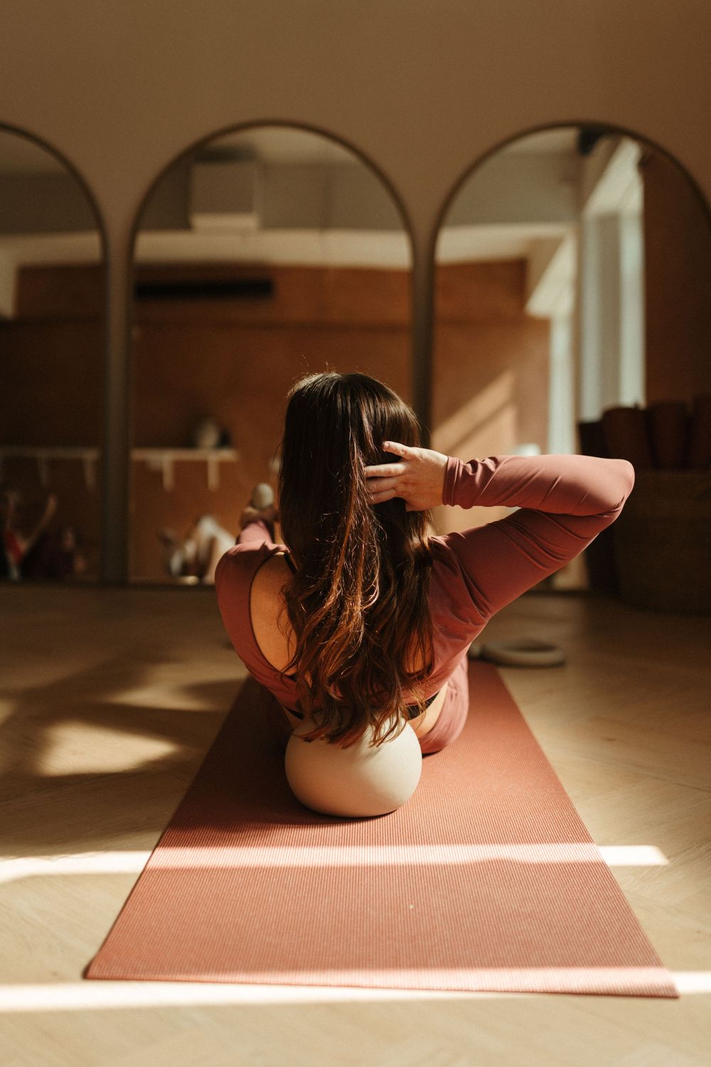 Woman practicing yoga on a red mat in a sunlit studio, sitting on a balance ball with hands behind her head.