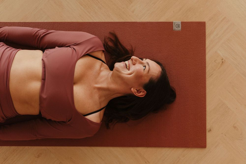 Woman in burgundy activewear lying on a yoga mat, smiling and relaxing during a workout session indoors.