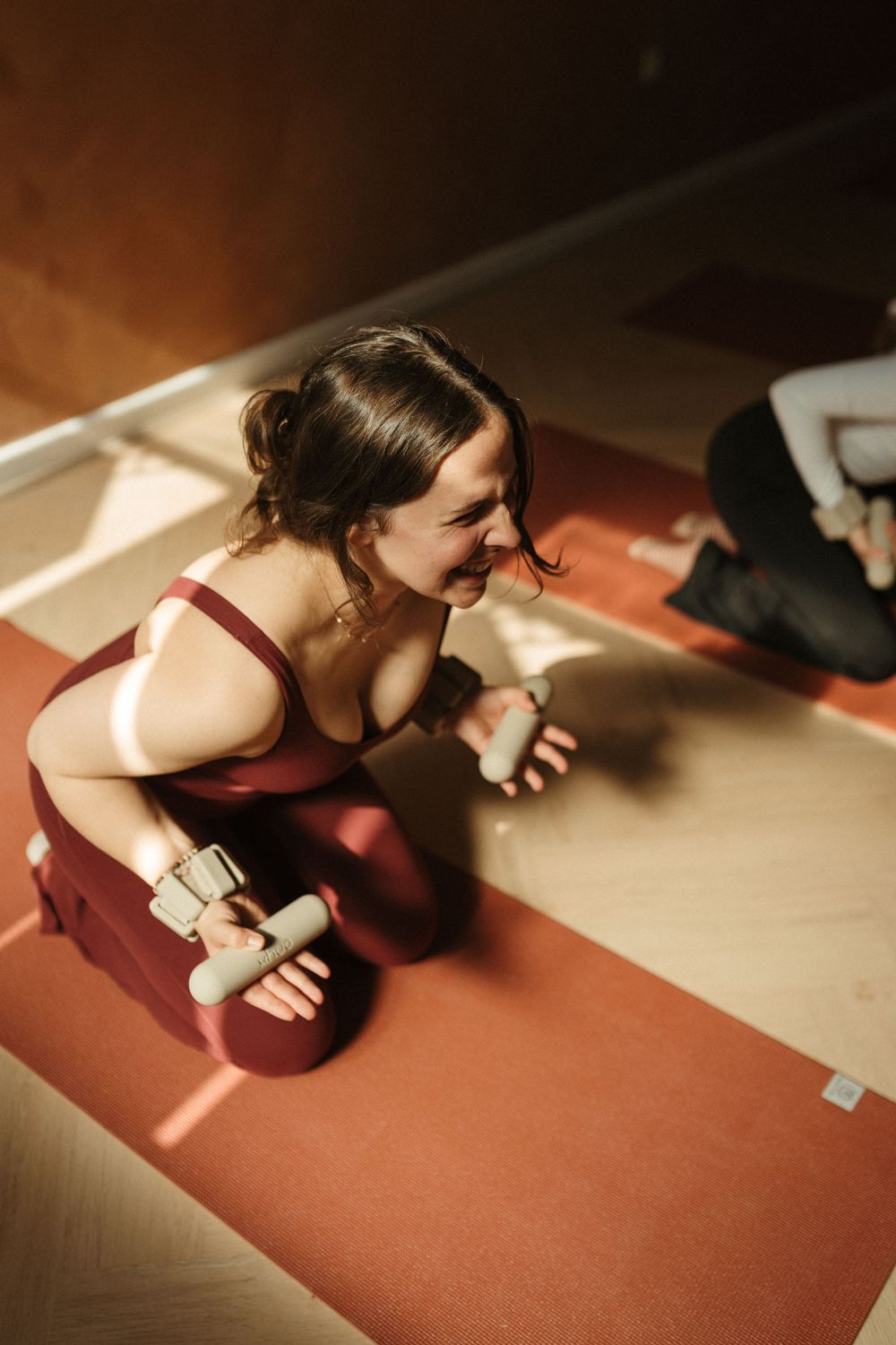 Smiling woman in burgundy workout top kneeling on yoga mat holding dumbbells during indoor fitness class.