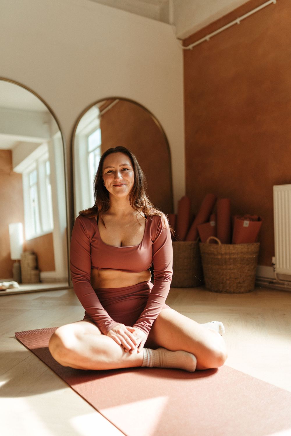 Woman in brown yoga outfit sitting cross-legged on a mat in a sunlit studio with arched mirrors and wicker basket.