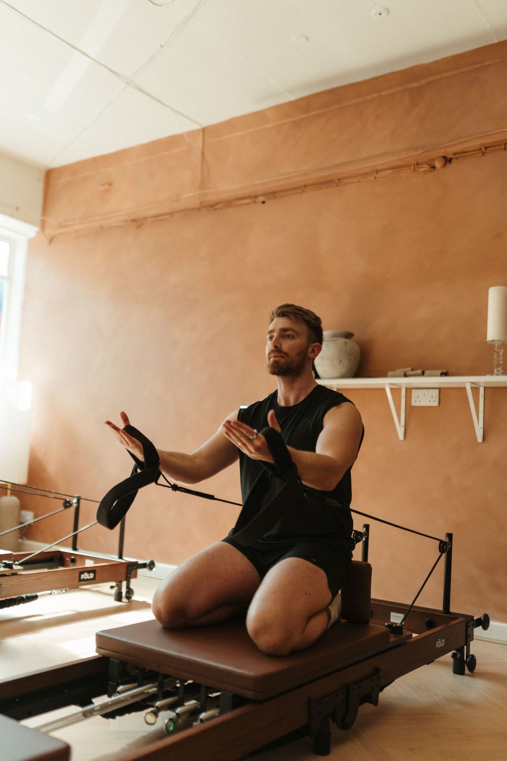 Man in black athletic wear kneeling on a Pilates reformer machine, clapping hands in a warm studio.