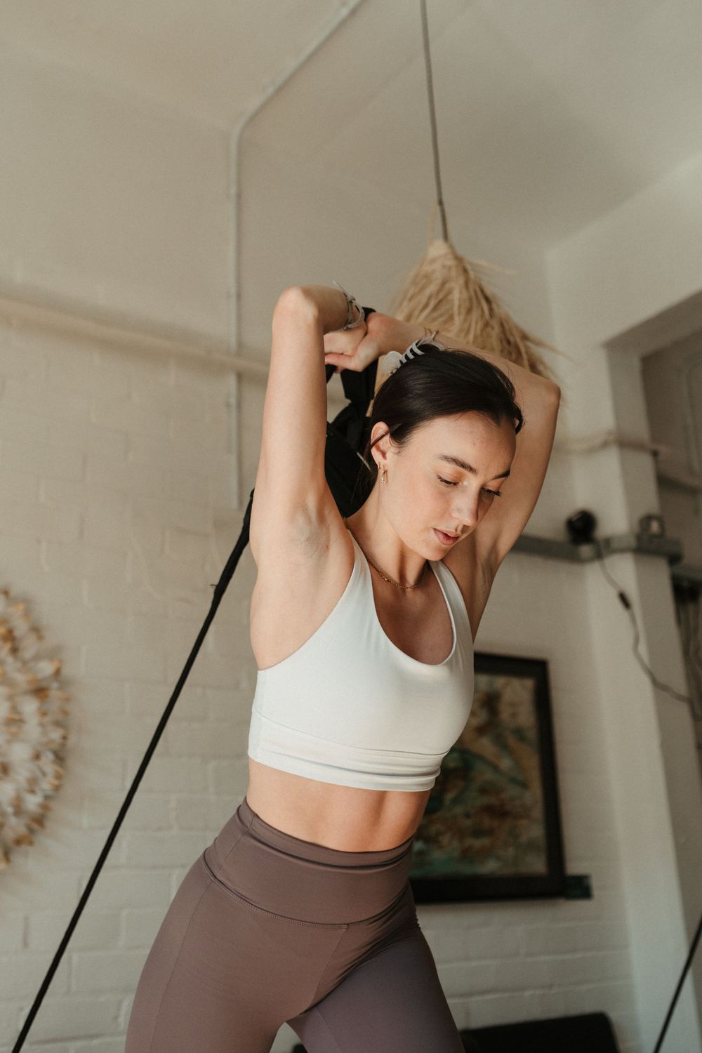 Woman in white sports bra and brown leggings stretching arms overhead during home workout session.