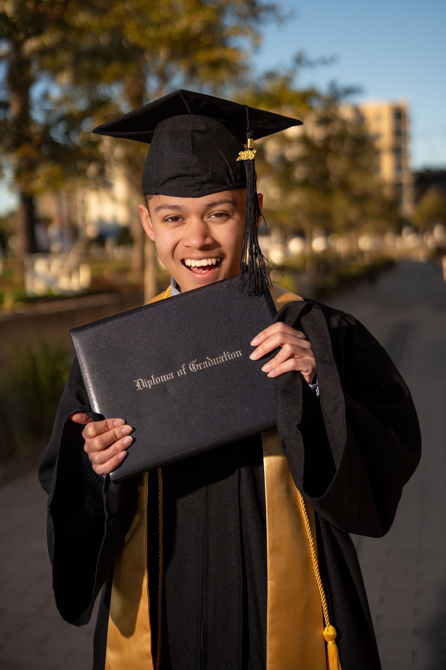 Smiling graduate in black cap and gown holds Diploma of Graduation outdoors in golden sunlight.