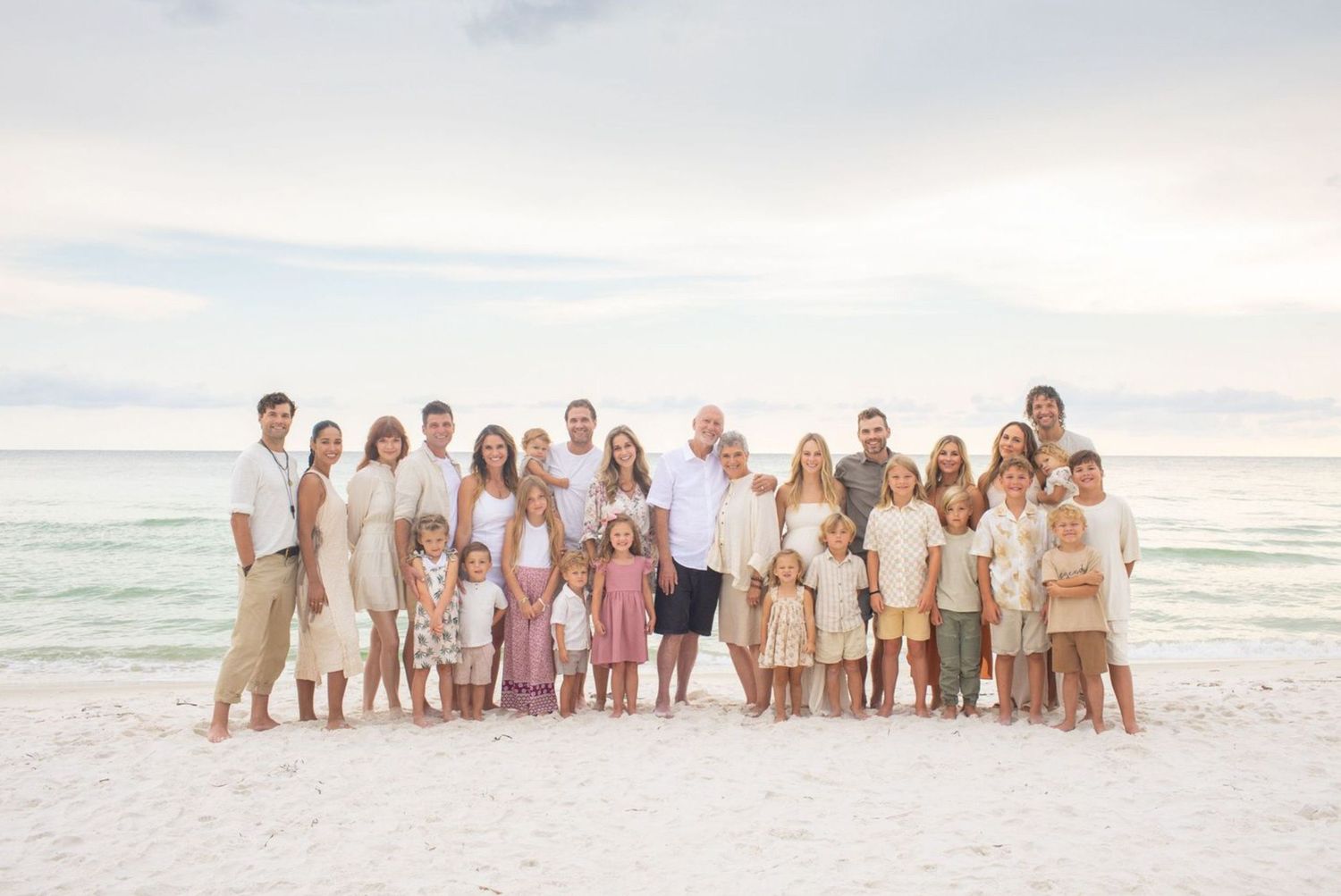 Large family group portrait on a white sand beach with calm turquoise water, wearing coordinated neutral and pastel outfits.