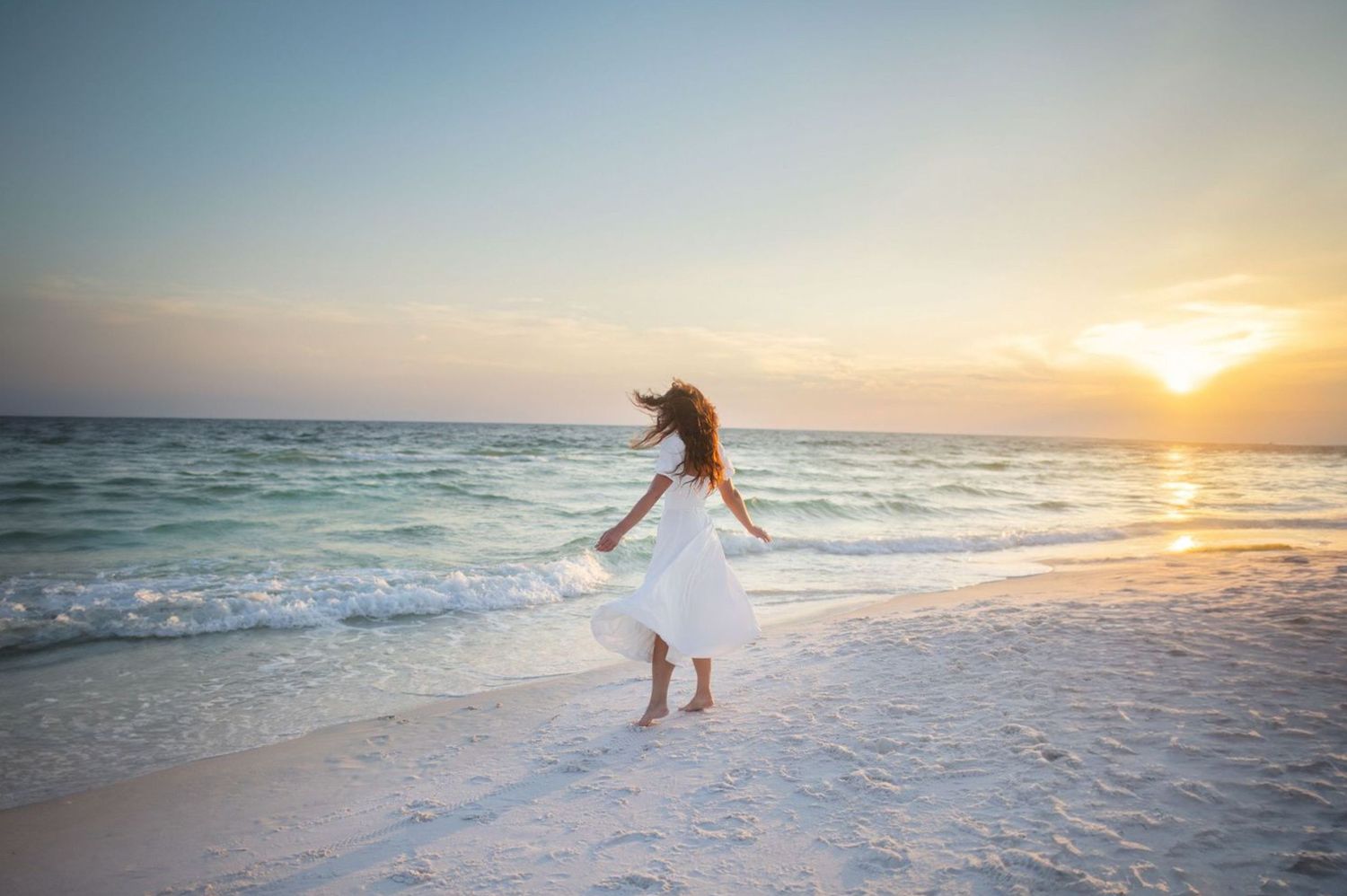 Woman in white dress and hat dancing on a white sand beach at golden sunset with calm ocean waves.