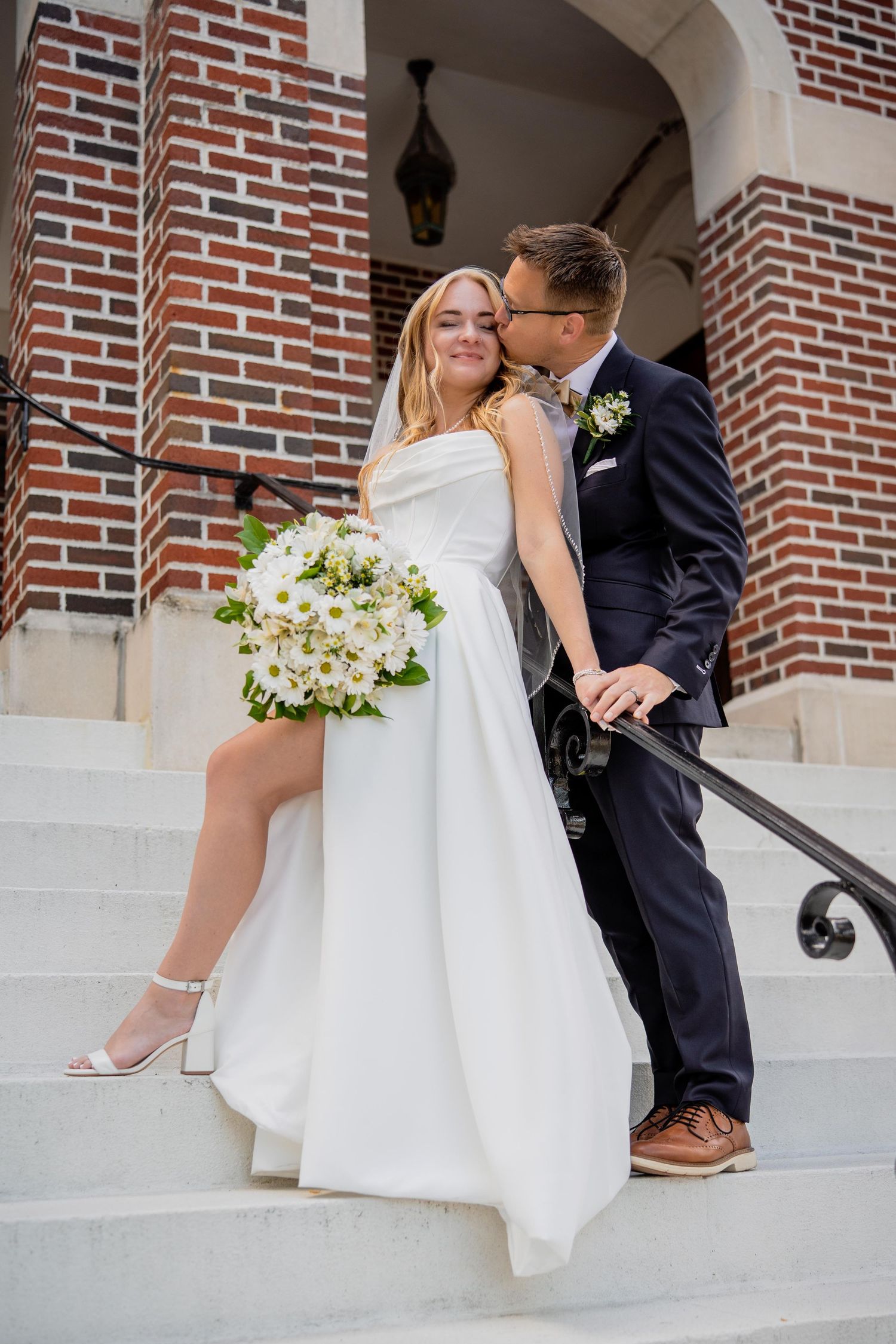 Bride in white gown holding daisy bouquet with groom in navy suit sharing a kiss on church steps.