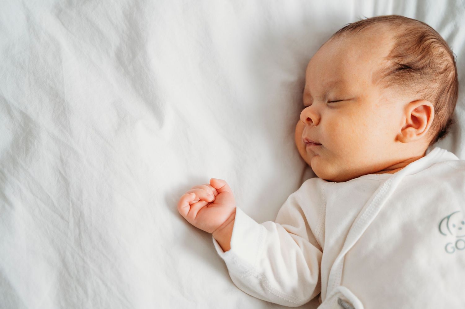 Newborn baby in white sleeper sleeps with hand out on white bed in Mississauga lifestyle newborn session