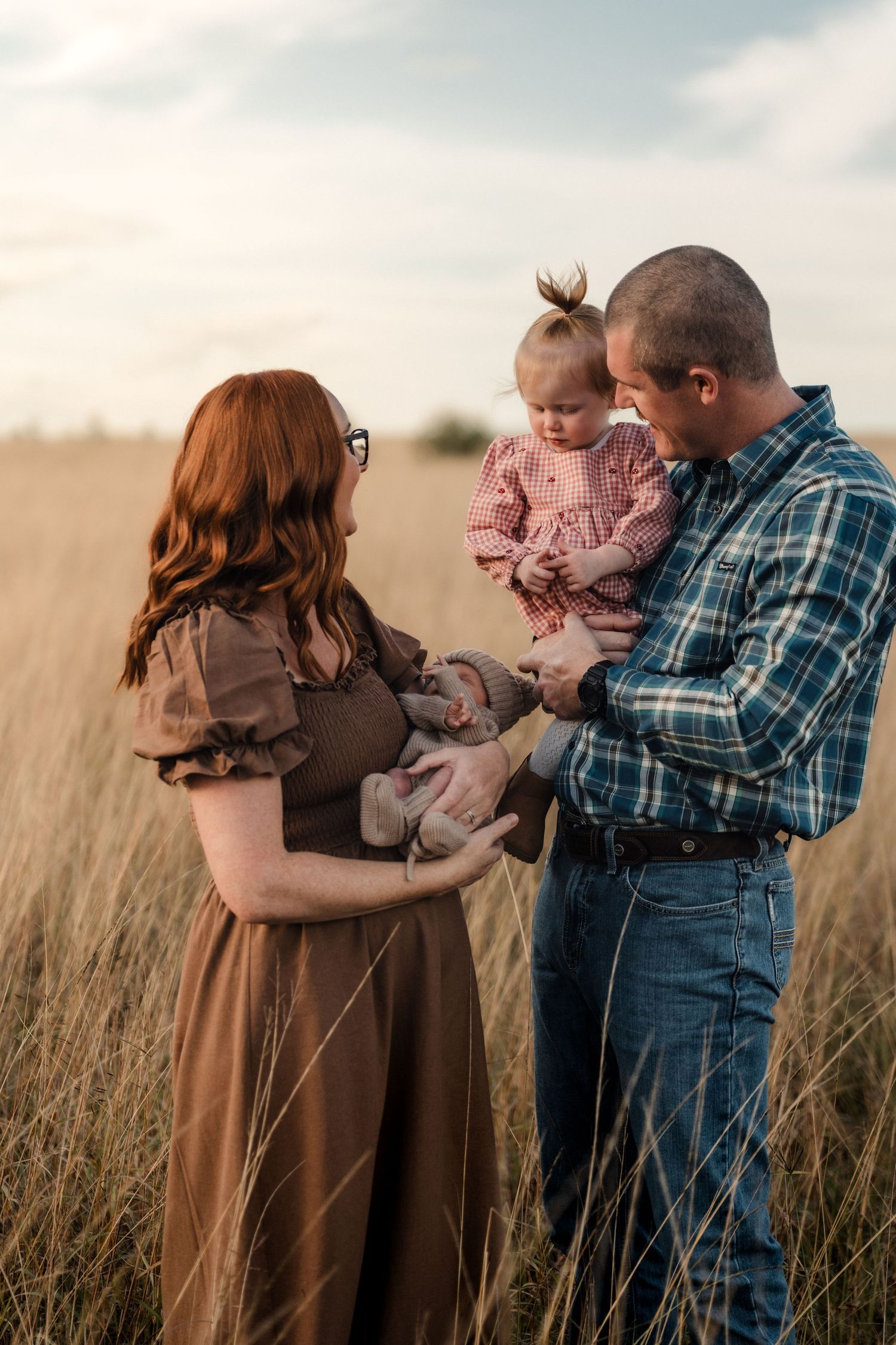 Tattooed blonde mother smiling lovingly at her newborn baby lying on a blanket in a golden grass field.