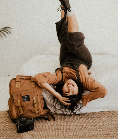 Woman lying upside down on a bed with a brown canvas backpack and camera beside her.