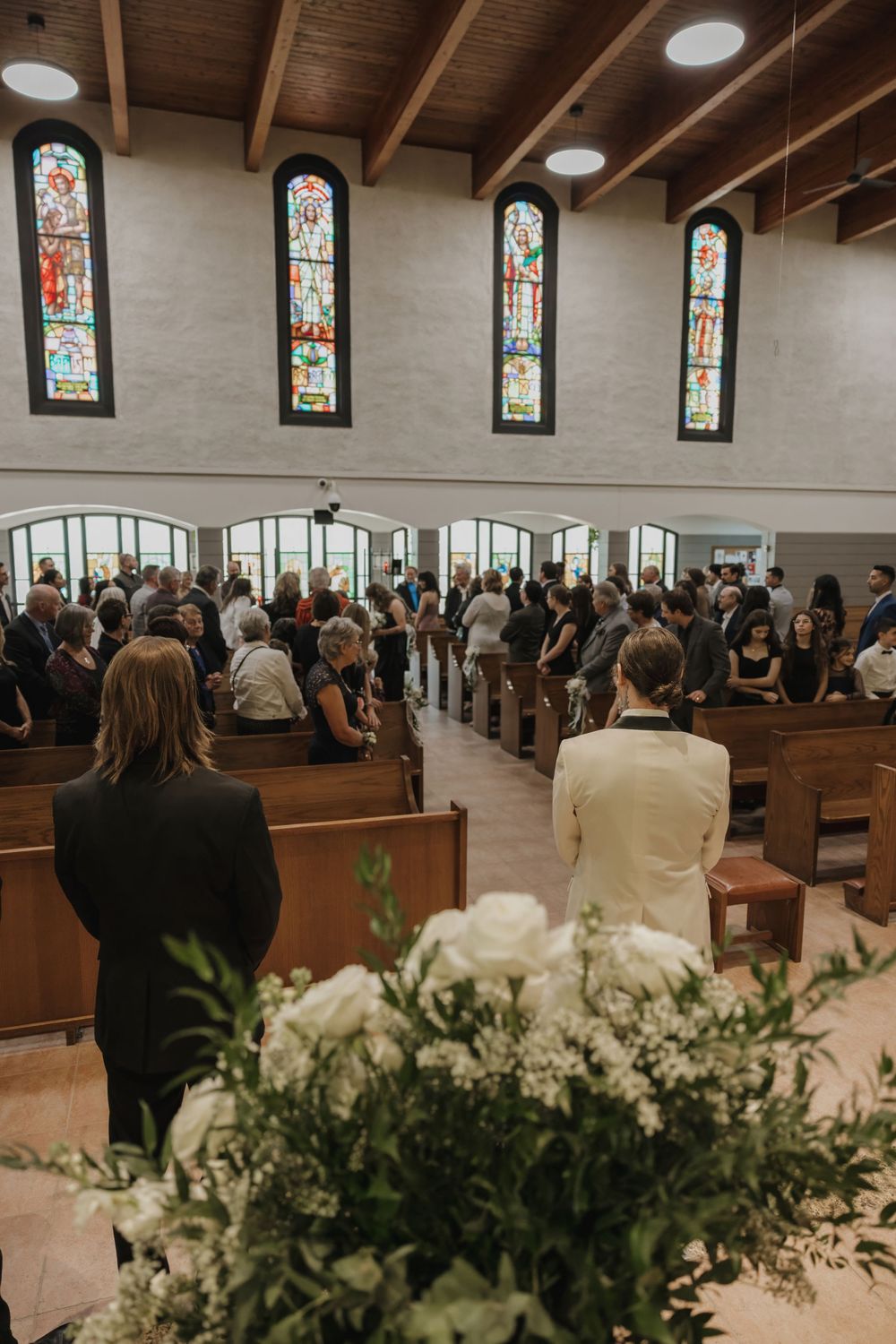 Wedding guests standing in a church aisle with stained glass windows and white floral arrangements.