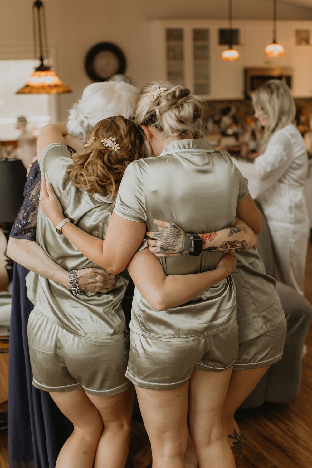 Bridesmaids in matching sage green satin pajama sets share a group hug during bridal preparations.