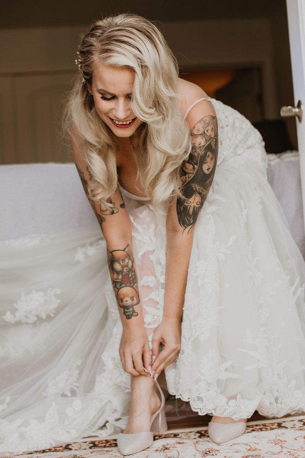Tattooed blonde bride in lace wedding gown smiling while fastening nude heels on wedding morning.