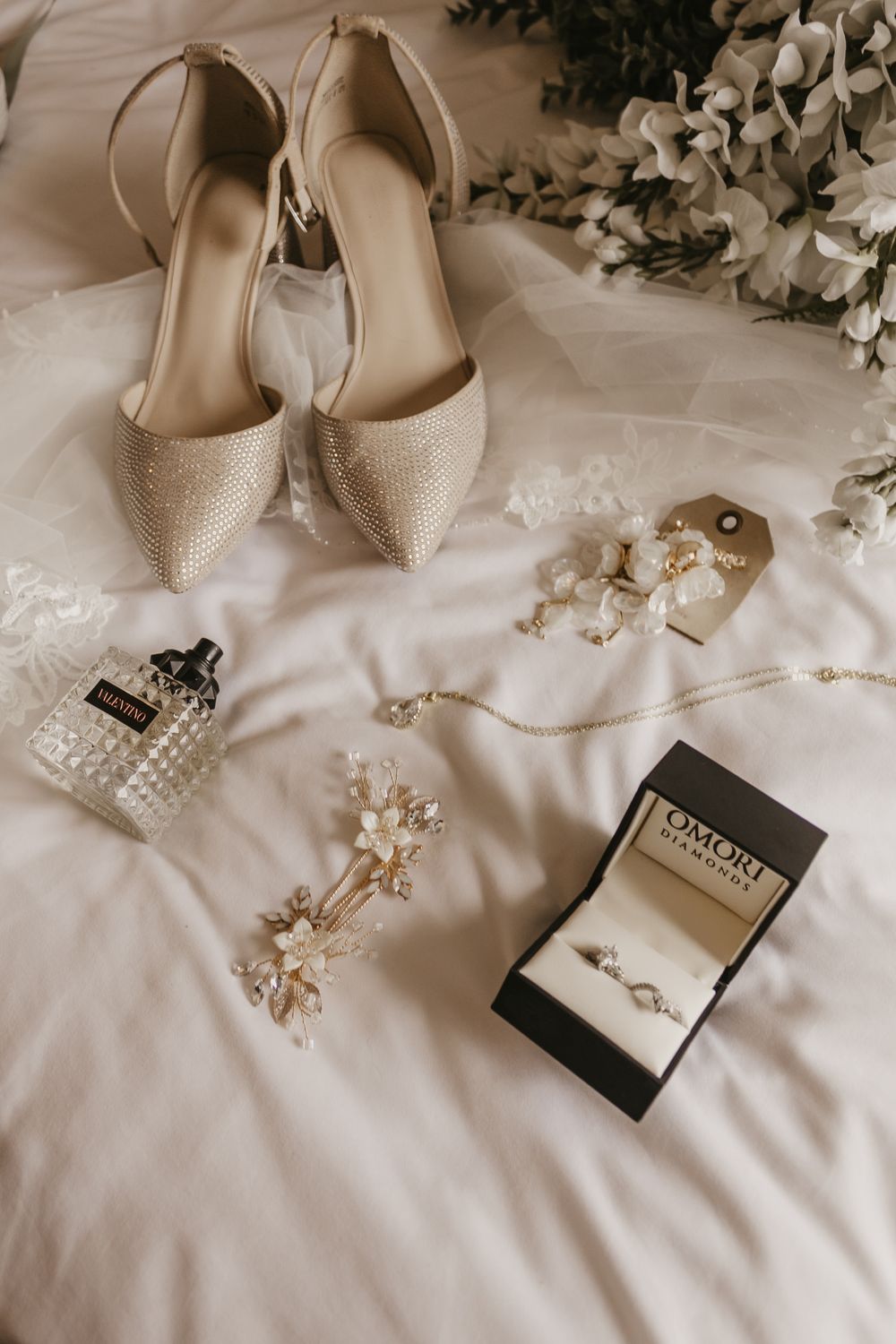 Bridal flat lay featuring nude heels, perfume, jewelry, and ring box on white bedding.